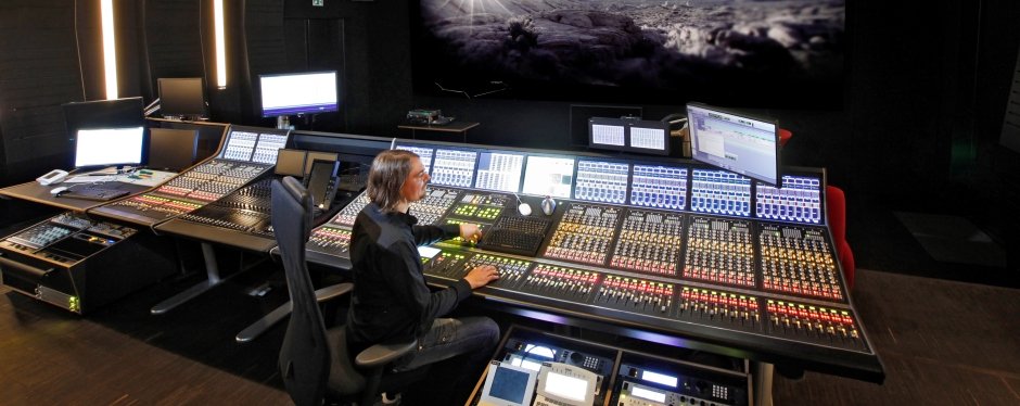 A man is sitting at a large illuminated mixing desk. In front of him there is a screen on which a black and white picture can be seen.