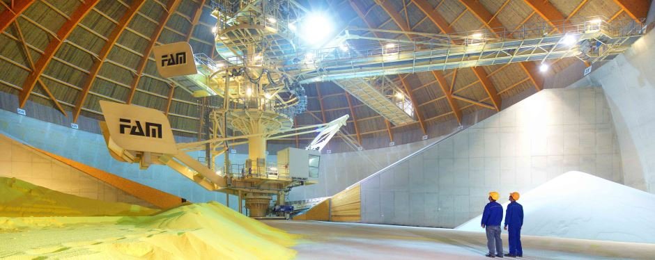 Two workers stand in a large factory hall. In the middle is a large conveyor system, in front of which is a layer of yellow sand-like material.